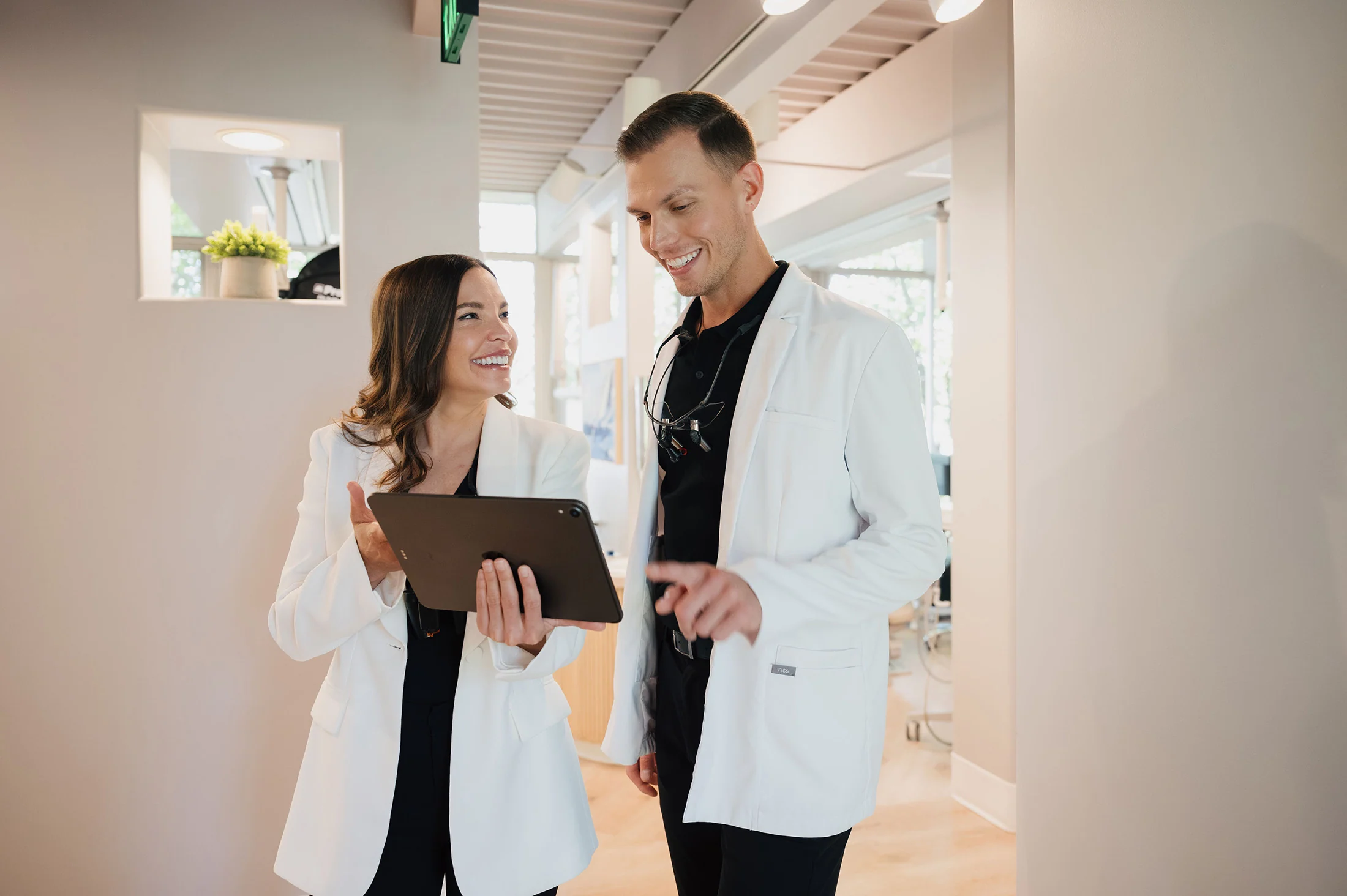 Two smiling medical professionals in white coats looking at a tablet in a bright office.