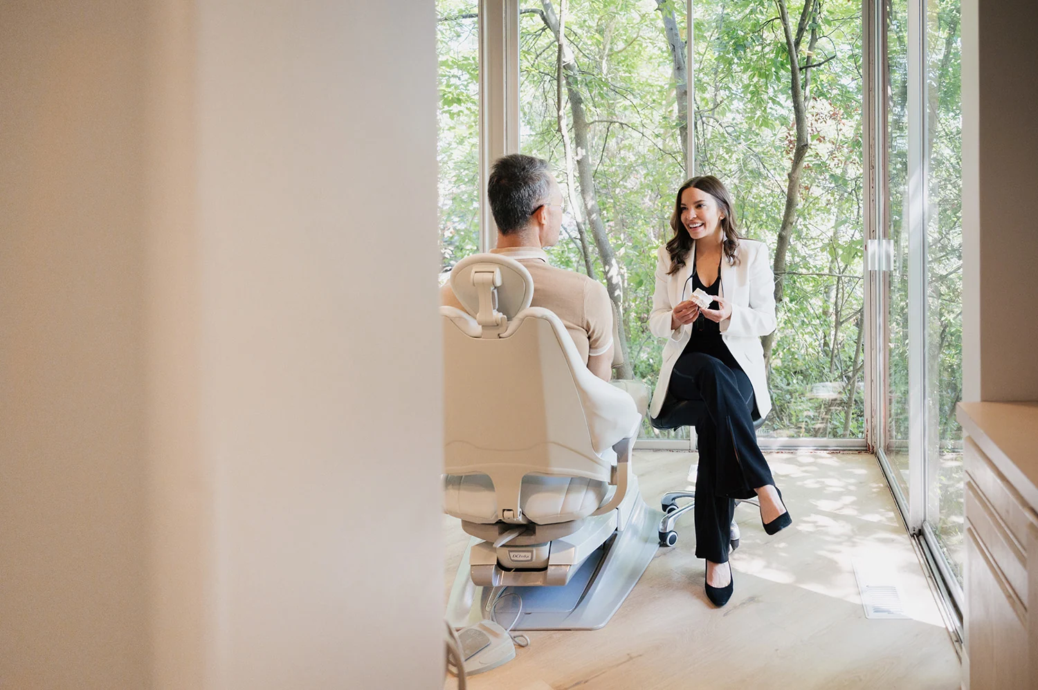 A woman in a white blazer smiling and talking to a man seated in a dental chair in a bright room with large windows showing green trees outside.